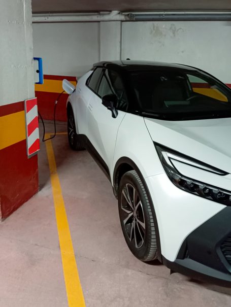 Coche blanco aparcado en un garaje, cerca de una pared y una barrera de acceso.